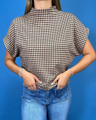 A woman wearing a brown and white gingham check short-sleeve blouse with a mock neck, paired with blue jeans, sta
