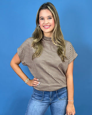 A woman wearing a brown and beige gingham short-sleeve mock neck top with blue jeans, posing against a solid blue background.