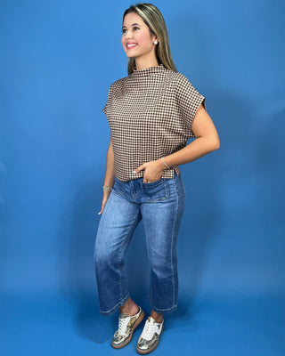 A woman wearing a brown and beige gingham short-sleeve mock neck top with blue jeans, posing against a solid blue background.