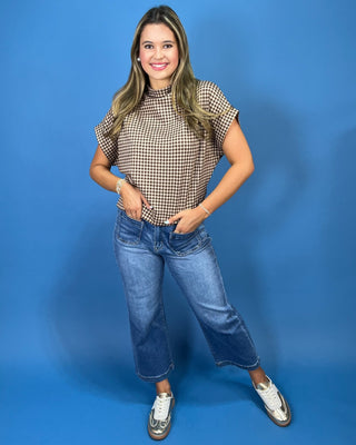 A woman wearing a brown and beige gingham short-sleeve mock neck top with blue jeans, posing against a solid blue background.