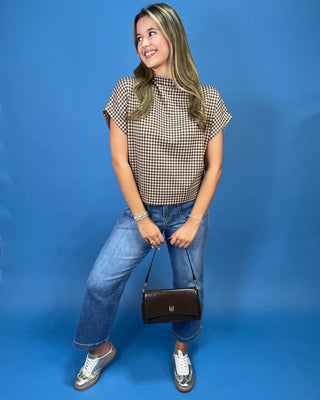 A woman posing in a brown and beige gingham short-sleeve top, blue jeans, and silver sneakers, holding a dark brown handbag a