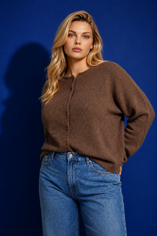 A woman modeling a brown fuzzy knit long-sleeve top with a drawstring neckline, paired with blue jeans, set against a solid dark blue studio background.