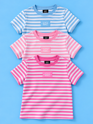 Three children's short-sleeve striped T-shirts from Lulu Mac are arranged vertically on a light blue background. The top shirt is light blue and white striped, the middle is pink and white striped, and the bottom is pink and light pink striped.