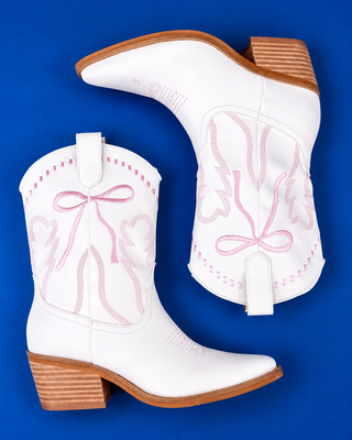 A pair of white leather cowboy boots with pink embroidered bow and leaf designs, displayed on a solid blue background.