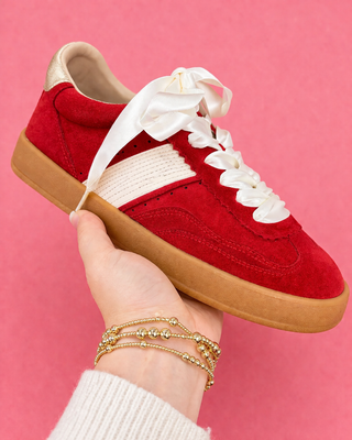 Red sneakers with white laces held by a hand against a pink background