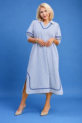 A woman modeling a blue and white vertically striped midi dress with blue piping details, standing against a solid blue studio background.