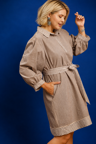 A woman modeling a taupe-brown linen-like shift dress with striped trim on the collar, cuffs, and hem, tied at the waist with a matching striped belt, set against a solid blue studio background.