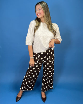 A woman modeling a light beige short-sleeve knit top and brown wide-leg pants with white polka dots, standing against a solid