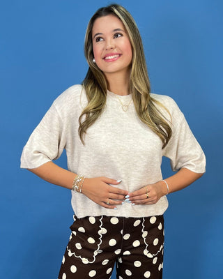 A woman modeling a cream-colored short-sleeve knit top and brown pants with white polka dots, standing a
