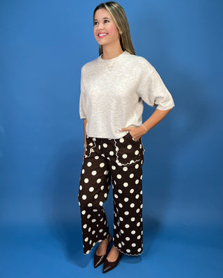 A woman modeling a light beige short-sleeve knit top and brown wide-leg pants with white polka dots, standing against a soli