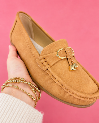 Brown loafer with gold buckle held by a hand against a pink background