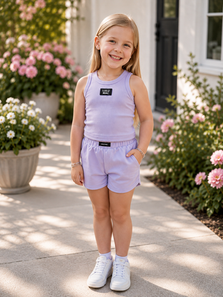A young girl stands outdoors on a sunlit walkway wearing a light lavender sleeveless tank top and matching shorts