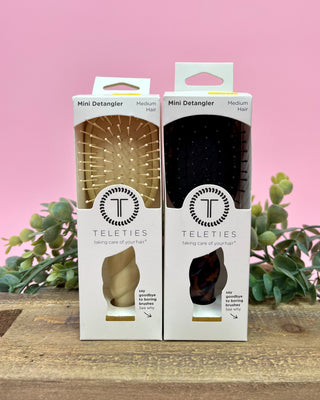 Two packages of Teleties Mini Detangler on a wooden surface with a pink background and plants.