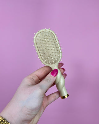 Hand holding a beige hairbrush with a gold accent against a pink background