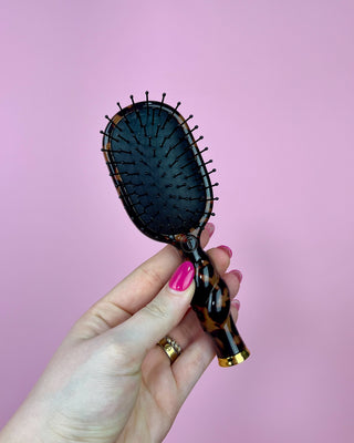Hand holding a tortoiseshell handle hairbrush against a pink background