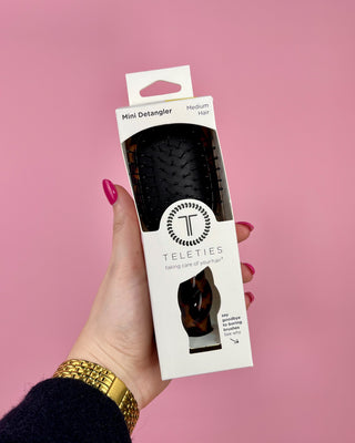 Hand holding a Teleties mini detangler packaging against a pink background