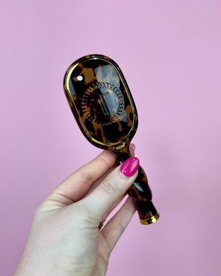 Hand holding a tortoiseshell-patterned hand mirror against a pink background
