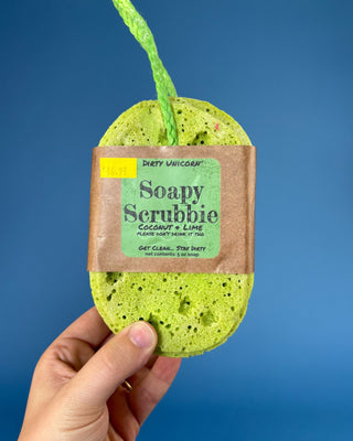 A hand holds a green porous scrub sponge with a brown paper label that reads 'Dirty Unicorn Soapy Scrubbie Coconut + Lime'
