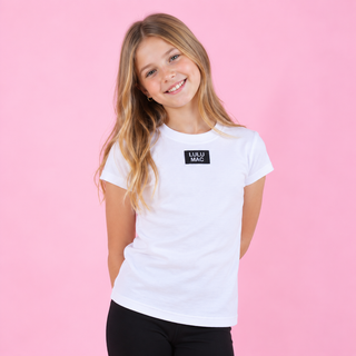 A young girl with long blonde hair smiling, wearing a white short-sleeve t-shirt with a black rectangular 'LULU MAC' logo patch on the chest, set against a solid pink background.