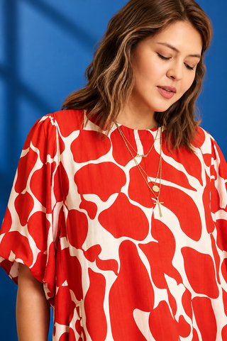 A woman wearing a red and off-white abstract patterned blouse with short puff sleeves, photographed against a blue studio background.