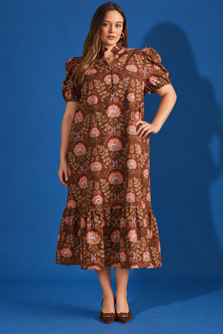 A woman modeling a brown bohemian-style midi dress with puff sleeves, a high ruffled collar, and an all-over floral print in pink, orange, and blue