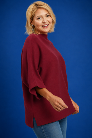 A woman with blonde hair wearing a loose-fitting, high-neck burgundy top and blue jeans, posing against a solid blue studio background.