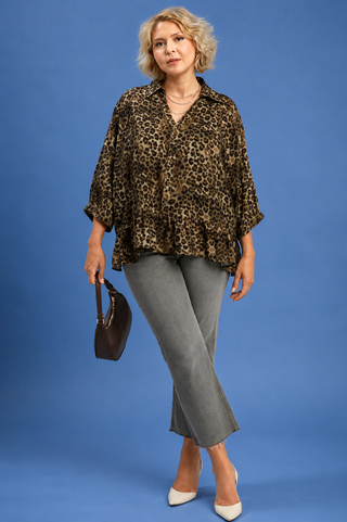 A woman modeling a leopard-print blouse with three-quarter sleeves, paired with gray cropped pants, white heels, and holding a dark brown handbag against a solid blue studio background.