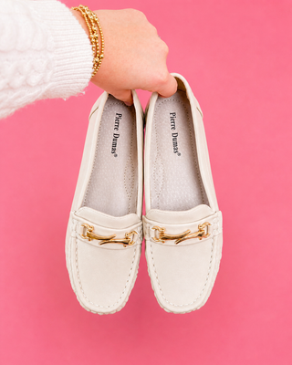 White loafers with gold accents held by a hand against a pink background