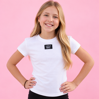 A young girl with blonde hair smiling and posing with her hands on her hips, wearing a white short-sleeve t-shirt with a black rectangular 'LULU MAC' logo patch on the chest, set against a solid pink background.