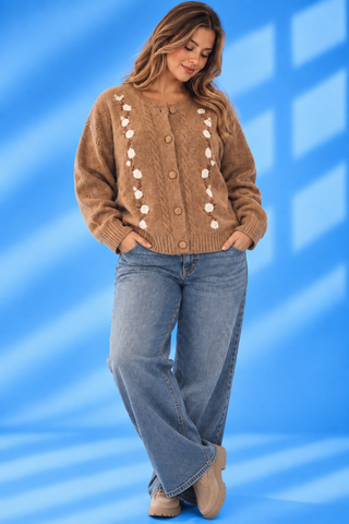 A woman modeling a brown knit cardigan sweater with white pom-pom accents down the front, paired with blue jeans and tan boots