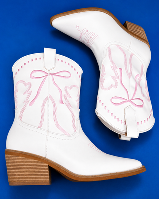 A pair of white leather cowboy boots with pink embroidered bow and heart designs, featuring brown stacked block heels, d