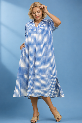 A woman modeling a light blue and white gingham midi dress with short sleeves, a V-neck, and side pockets, posed against a solid blue studio background.