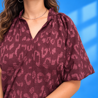 A close-up of a woman wearing a short-sleeve crinkled blouse with a dusty rose and burgundy abstract print, paired with a silver chain necklace,
