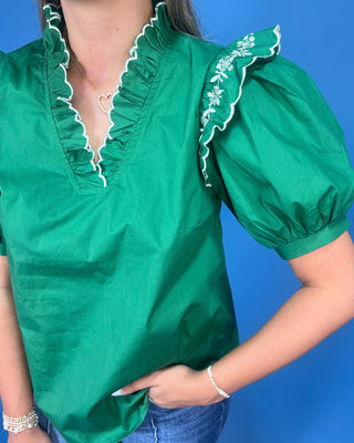 A green short-sleeve blouse with white ruffle trim and white floral embroidery on the shoulder, worn by a model against a