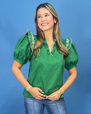 A woman wearing a bright green short-sleeve blouse with white embroidered ruffle trim at the collar ana
