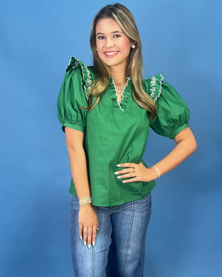 A woman modeling a green puff-sleeve blouse with white lace trim at the ruffled neckline, paired with blue jeans, against a s