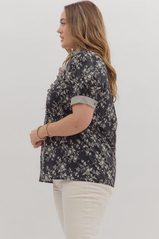 Woman wearing a black floral blouse with white pants on a plain background