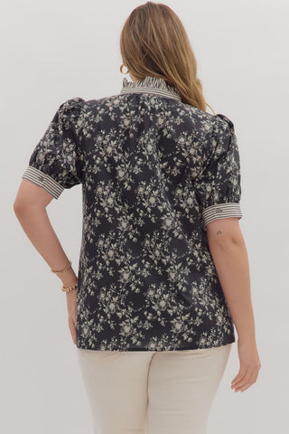 Woman wearing a black floral blouse with white pants on a plain background