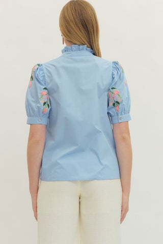 Light blue blouse with floral embroidery on sleeves worn by a person against a white background