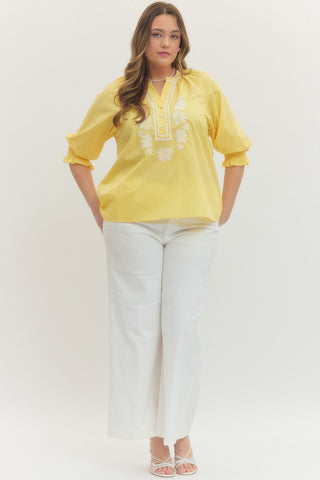 Woman wearing a yellow embroidered blouse and white pants on a plain background