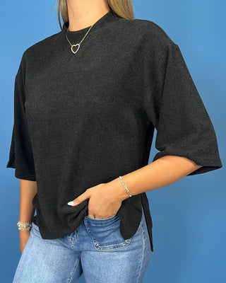 A woman wearing a dark charcoal gray oversized short-sleeve top with a side slit, paired with light blue jeans, standing agai