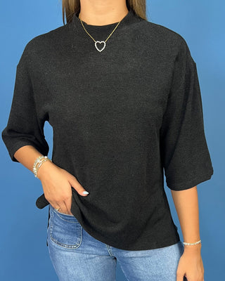 A person wearing a dark charcoal gray knit three-quarter sleeve top with a high mock neck, paired with blue jeans, standing a