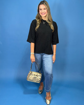 A woman modeling a black short-sleeve top, blue jeans, and metallic gold shoes, holding a metallic gold handbag against a sol