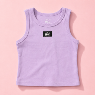 A young girl stands outdoors on a sunlit walkway wearing a light lavender sleeveless tank top and matching shorts