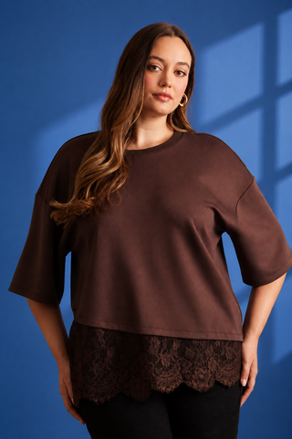 A woman modeling a brown short-sleeve top with a scalloped lace hem, posed against a blue background with window-pane shadows.