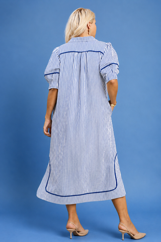 A woman viewed from the back wearing a blue and white vertically striped midi dress with dark blue piping details, standing against a solid blue studio background.