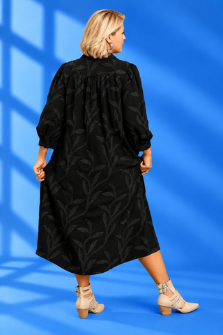 A woman modeling a black midi-length collared dress with a subtle tonal leaf pattern