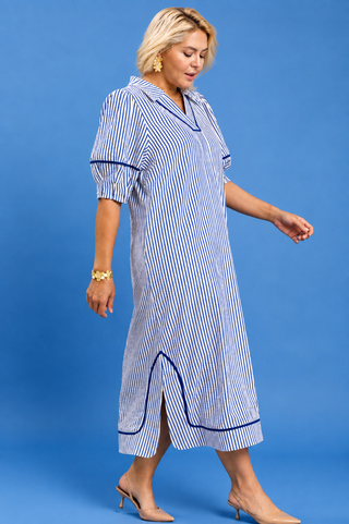 A woman modeling a blue and white vertically striped midi dress with dark blue piping details, standing against a solid blue studio background.