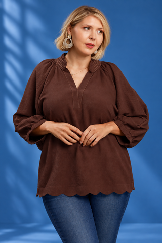 A woman modeling a brown long-sleeve blouse with a scalloped hem and gathered neckline, paired with blue jeans, against a blue studio backdrop with soft shadow overlays.