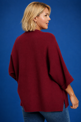 A woman with blonde hair is shown from the back wearing a loose-fitting, deep burgundy poncho-style top and blue jeans against a solid blue studio background.
